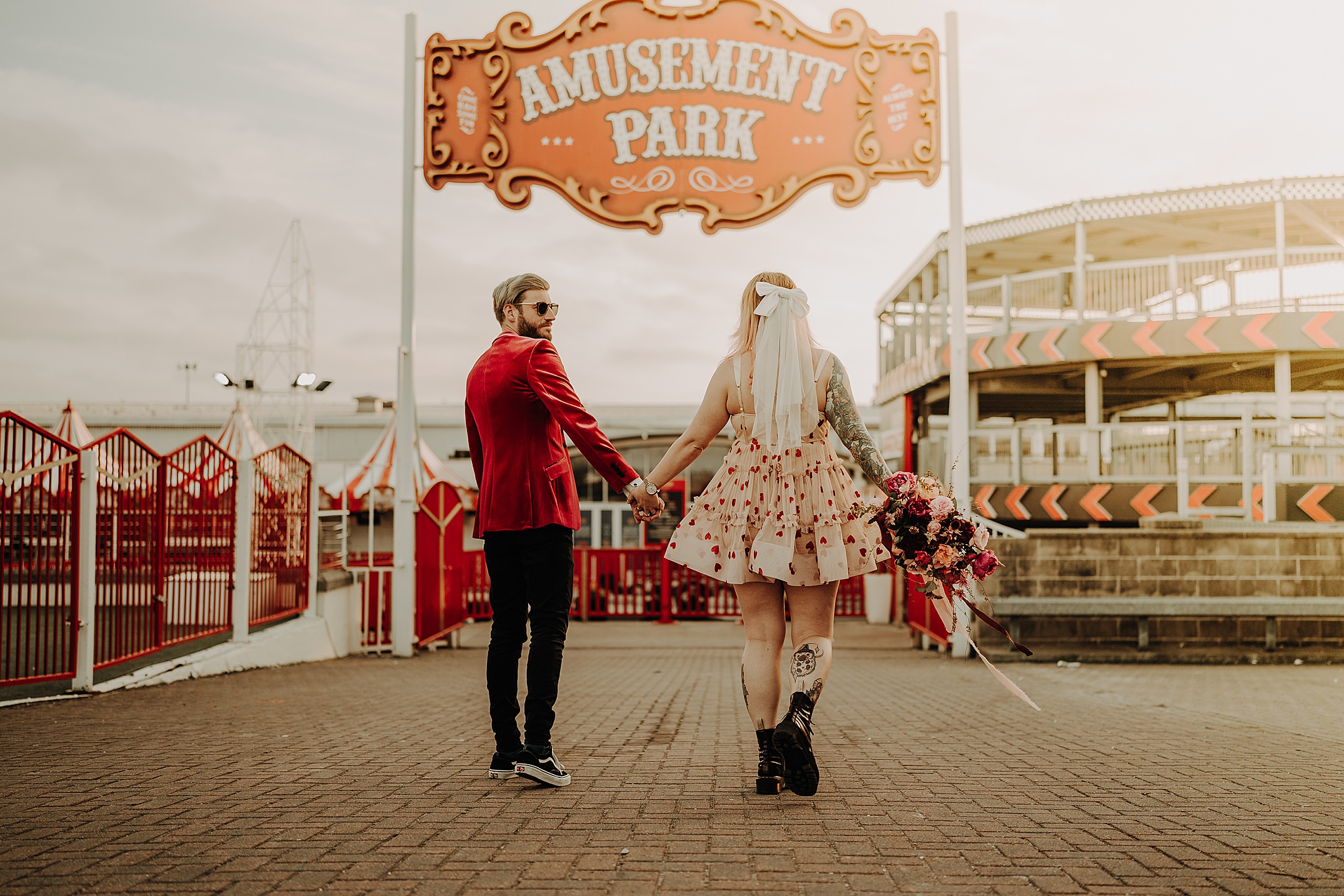 couple hand in hand walking away from camera into amusement park she is wearing dress with love hearts and he is wearing red velvet jacket scotland engagement photos aberdeen