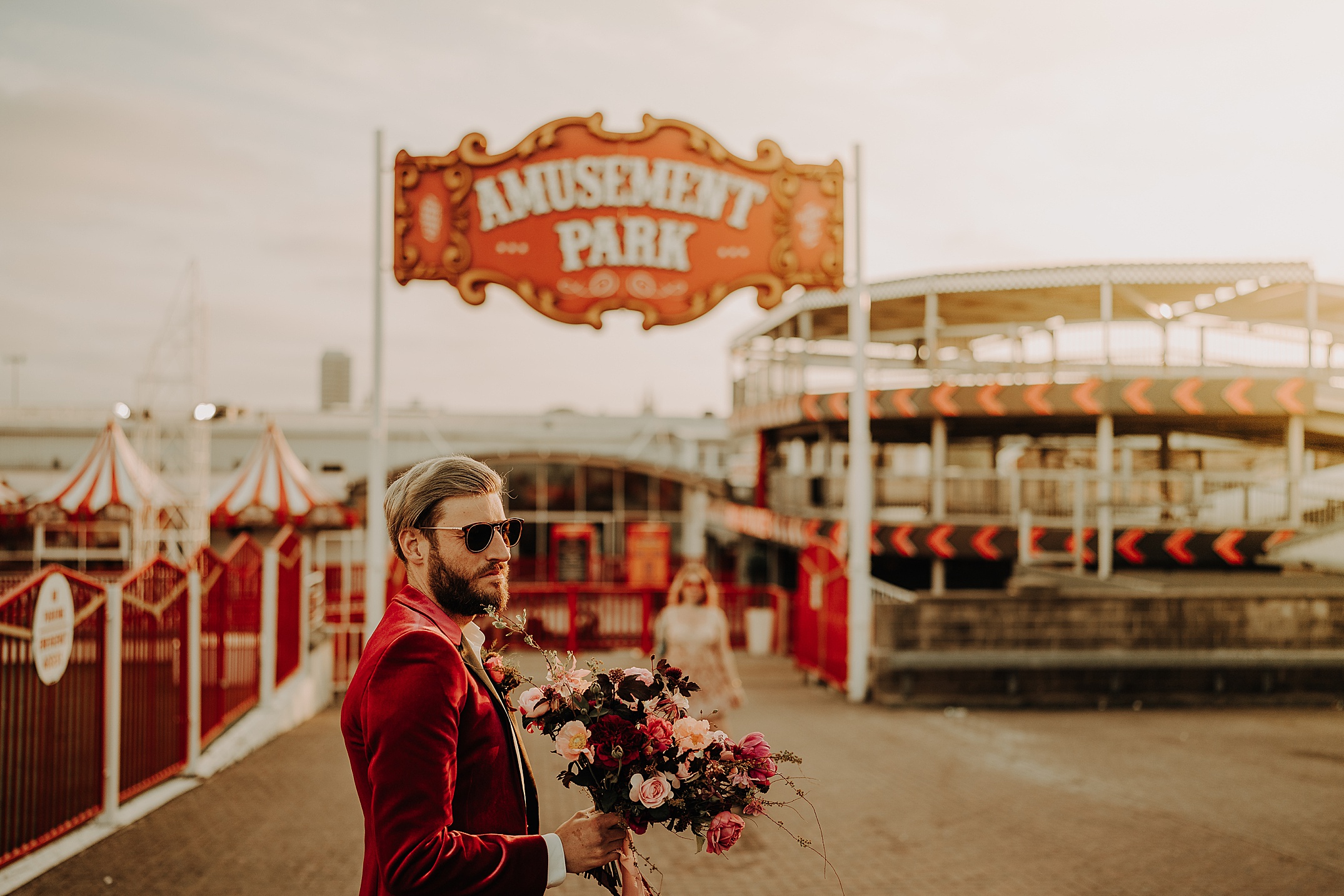 guy wearing red vevert jacket holding bouquet of flowers in front of amusement park sign as she walks towards him scotland engagement photos aberdeen
