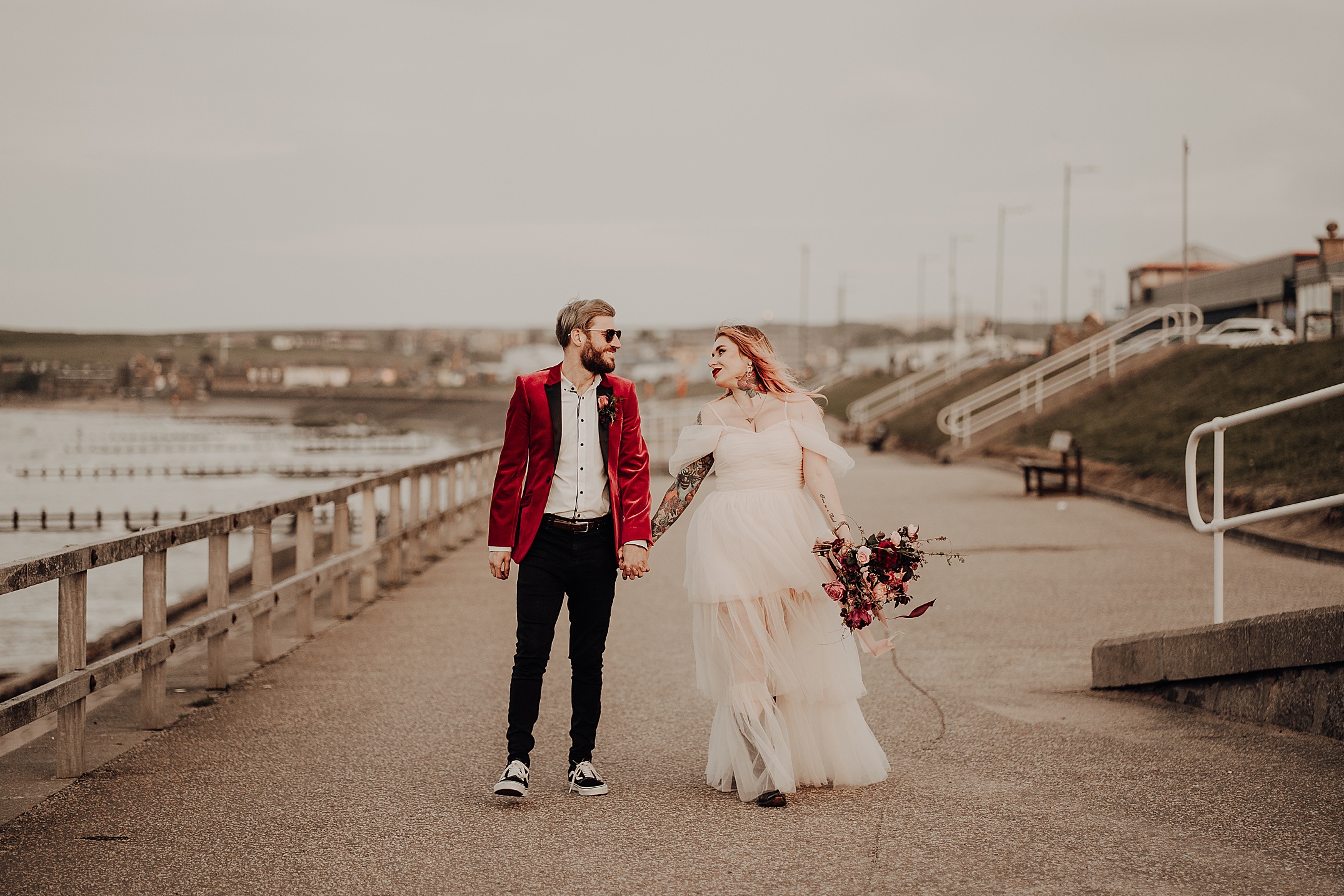 she is wearing white dress and holding bouquet he is wearing red velvet jackets as they hold hands walking down aberdeen promenade scotland engagement photos