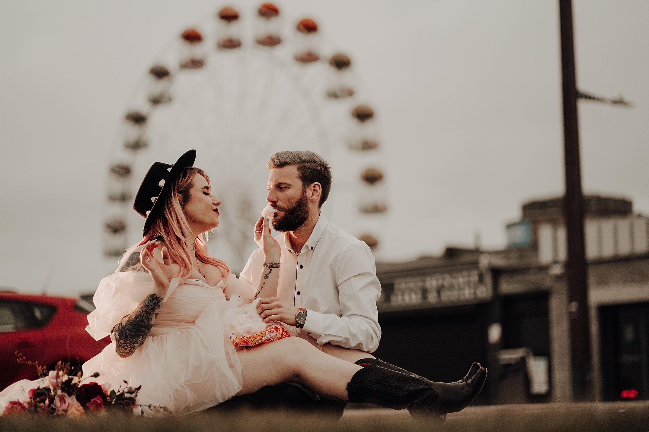 girl wearing white dress black boots and black hat feeding him candy floss with big wheel in background scotland engagement photos aberdeen