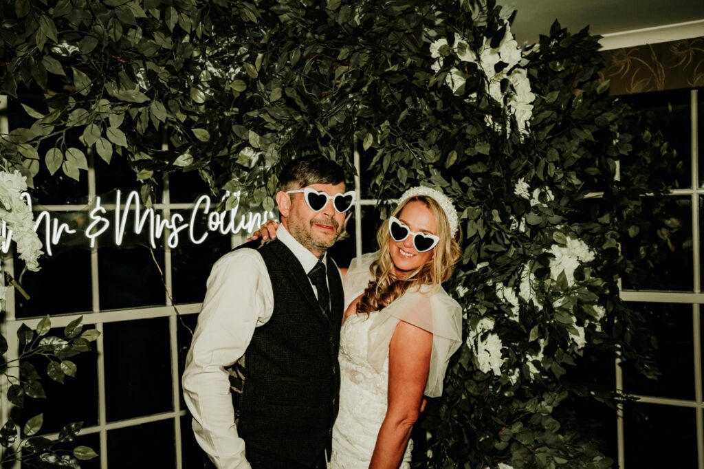 Couple in front of greenery garland and neon sign, wearing heart shaped glasses at Banchory Lodge Hotel