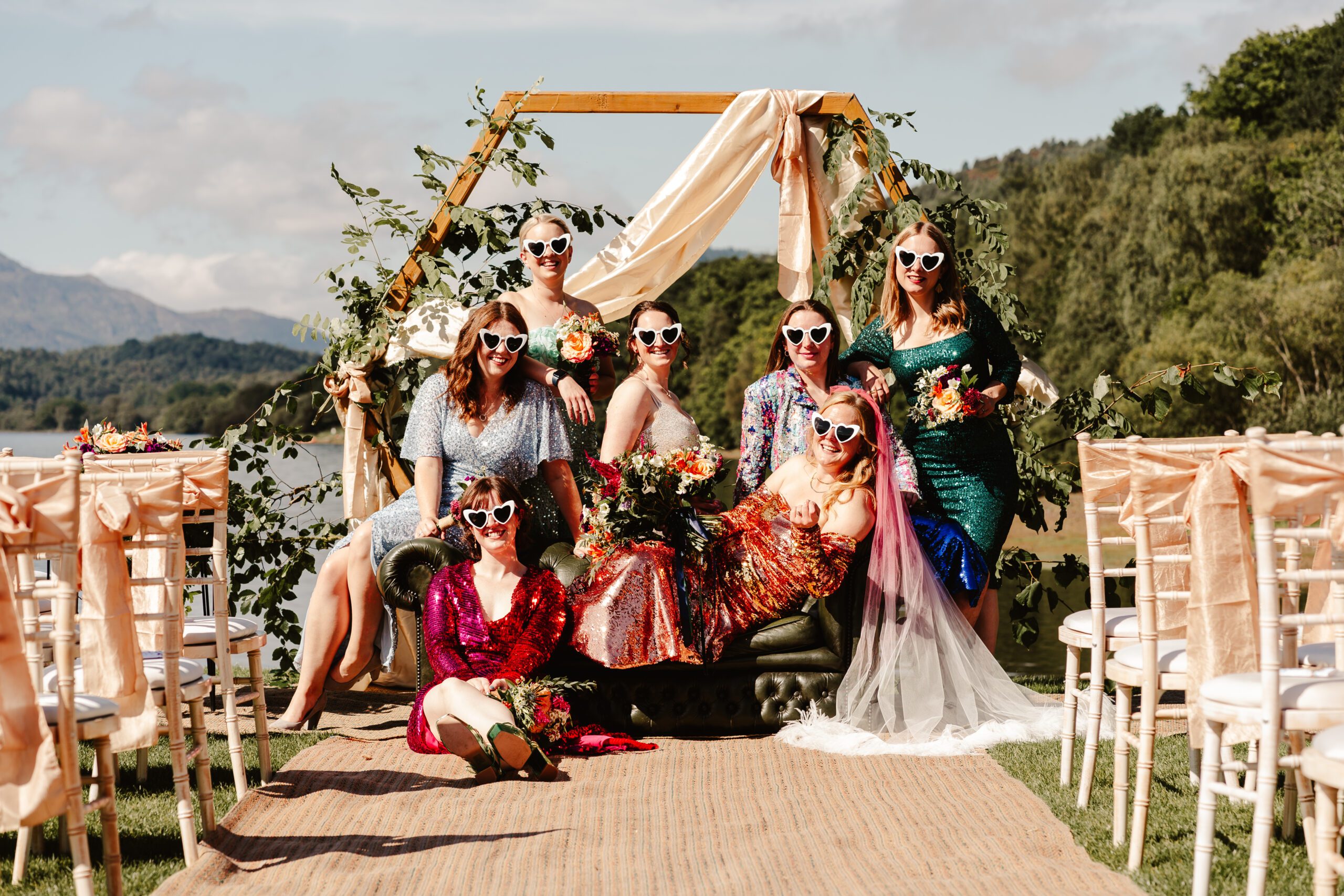 Bridal party wearing sequin dresses pose on sofa at scottish outdoor wedding wearing heart shaped sunglasses