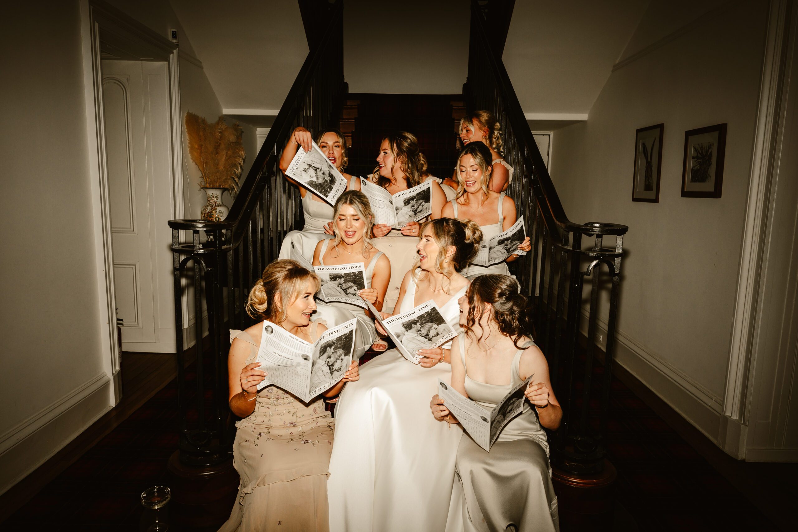 Bridal party sit on stairs reading wedding newspaper at netherdale house